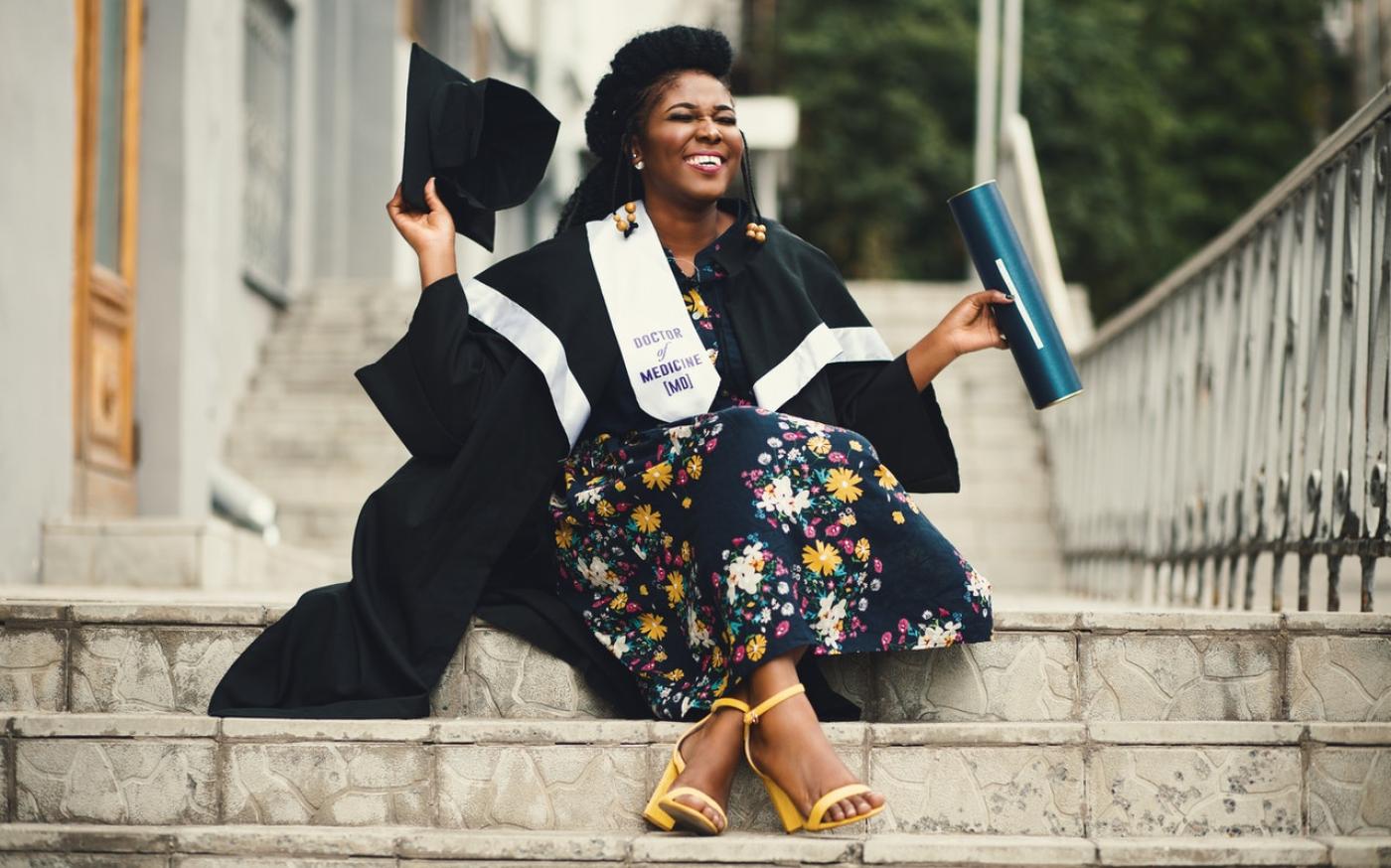 Woman in grad cap and gown sitting on stairs and smiling. She's wearing a ribbon that says Doctor of Medicine